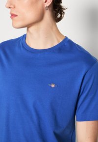 Modré bavlněné tričko s kulatým výstřihem, se malým vyšitým logem na hrudi. Hladký povrch a krátké rukávy.