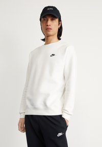 Valkoinen sweatshirt, jossa on pyöreä kaula-aukko ja pitkät hihat, pienen mustan Nike-logon kanssa rinnassa, yhdistettynä mustiin verkkohousuihin. Musta lippalakki.