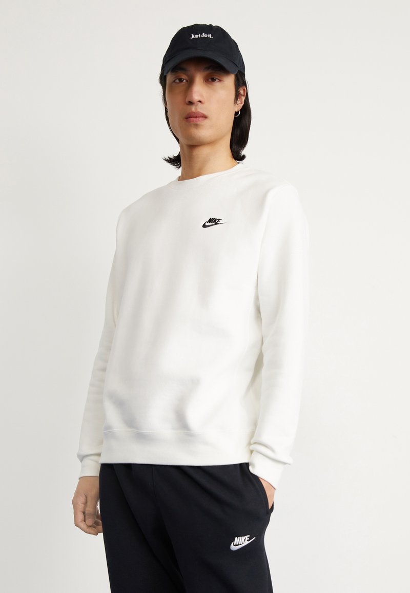 Valkoinen sweatshirt, jossa on pyöreä kaula-aukko ja pitkät hihat, pienen mustan Nike-logon kanssa rinnassa, yhdistettynä mustiin verkkohousuihin. Musta lippalakki.