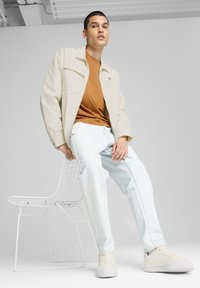 Beige dzseki egy barna póló felett, világoskék farmer, és fehér edzőcipő. Fehér drótszéken ül, egy sima háttér előtt.