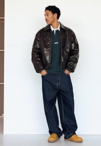 Man wearing dark brown leather jacket, green collared shirt, loose dark jeans, and tan boots, standing with hands in pockets looking sideways.