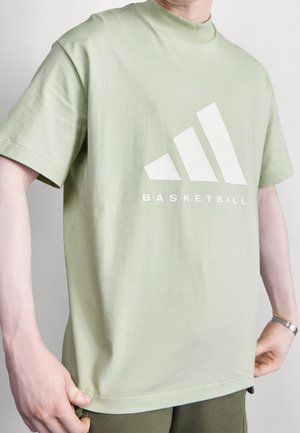 Mann trägt ein hellgrünes T-Shirt mit weißem stilisiertem Logo und dem Wort "BASKETBALL" auf der Vorderseite, Hände, die den Saum des Shirts anpassen.