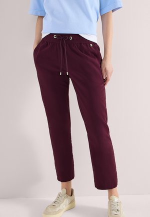 Osoba nosząca bordowe spodnie ze sznurkiem, jasnoniebieską bluzkę, biały podkoszulek oraz beżowe tenisówki, stojąca na jednolitym tle.