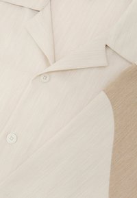 Blazers pliés crème et beige avec revers crantés et deux boutons couleur crème sur une surface en tissu texturé.