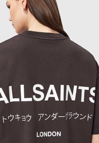 Σκούρο καφέ υπερμεγέθη μπλουζάκι, βαμβακερό ύφασμα, με μεγάλα λευκά γράμματα στο πίσω μέρος: "ALLSAINTS" και άλλα κείμενα σε διάφορες γλώσσες.