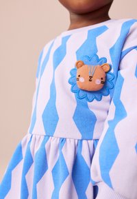 Plava i bijela prugasta haljina s nabranim suknjom, s aplikacijom svezena lava u obliku plavog cvijeta na sredini sprijeda.