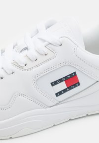Zapatilla blanca de cuero con superficie texturizada, parte delantera con cordones, cuello acolchado y un parche con logotipo que presenta acentos rojos y azules en el lateral.