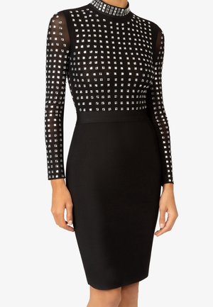 Robe noire moulante avec manches longues transparentes et haut orné de petits clous carrés argentés disposés en motif quadrillé.