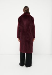 Burgundy faux fur coat with a collar, long sleeves, a central seam, and a back slit, styled over black wide-leg trousers.