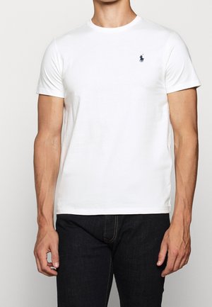Homme portant un t-shirt blanc uni à manches courtes avec un petit logo Polo foncé sur la poitrine gauche et un jean foncé.
