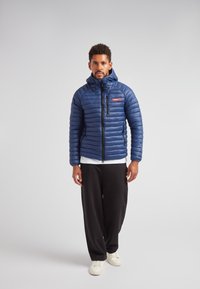 Marinblå pufferjacka med horisontellt quiltat mönster, framdragkedja och huva. Buren över en vit t-shirt och svarta byxor.