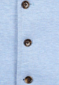 Helles blaues, strukturiertes Gewebe mit einem karierten Muster, das vier dunkelbraune Knöpfe auf der Vorderseite aufweist. Maschinengenähte Knopflöcher.