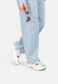 Zapatillas multicolores con acentos en blanco, verde, rosa y azul, combinadas con vaqueros de pierna ancha de color azul claro. Gafas sujetas con una mano.