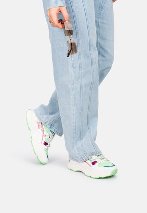 Zapatillas multicolores con acentos en blanco, verde, rosa y azul, combinadas con vaqueros de pierna ancha de color azul claro. Gafas sujetas con una mano.