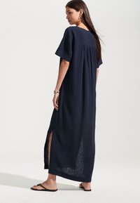 Tumši zila lina maxi kleita ar īsām piedurknēm, aizmugurē krokojumu un sānu šķēlumiem; teksturēts audums, vaļīga piegrieztne, valkāta ar melnām flipflop sandales.