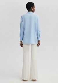 Camisa de manga larga azul claro con cuello y puños abotonados, combinada con pantalones de campana blancos de talle alto y un acabado texturizado.