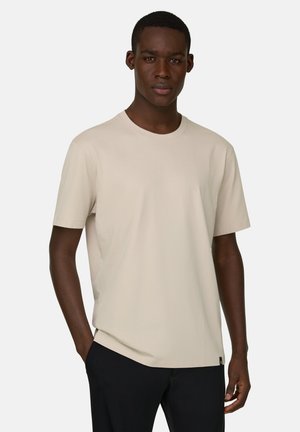 Hombre joven vestido con una camiseta beige lisa de manga corta y pantalones negros, de pie con una mano en el bolsillo contra un fondo claro.
