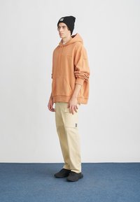Sweatshirt laranja com um bolso canguru e letras pretas, combinado com calças de carga bege. Um gorro preto e sapatilhas pretas completam o visual.
