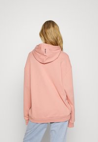 Sudadera con capucha rosa de textura suave, ajuste oversized y dobladillo acanalado. Presenta un logo en la parte posterior, cerca del cuello. Combinada con pantalones de color azul claro.