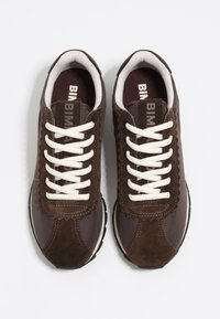 Un par de zapatillas oscuras marrones con cordones blancos y un acolchado interior blanco sobre un fondo blanco.