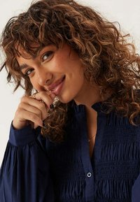 Vrouw met krullend bruin haar, gekleed in een marineblauw blouse, glimlacht zachtjes en rust haar hand bij haar gezicht, met een ring om haar vinger.