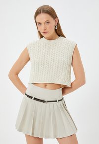 Un suéter sin mangas corto de estilo crochet en color crema claro, combinado con una falda beige con pliegues y de cintura alta, acentuada con un cinturón negro.