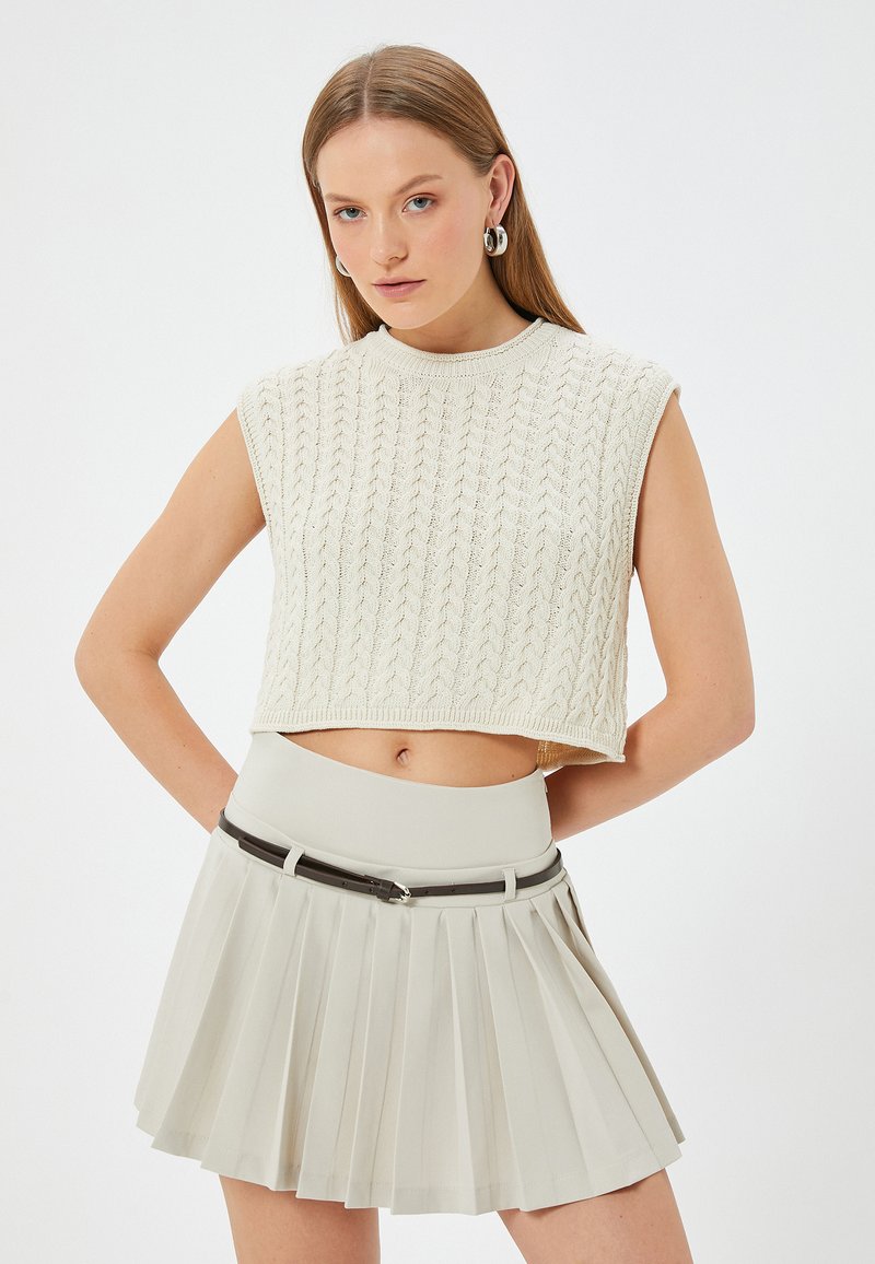 Un suéter sin mangas corto de estilo crochet en color crema claro, combinado con una falda beige con pliegues y de cintura alta, acentuada con un cinturón negro.