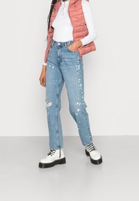 Vadderad rosa väst över en vit långärmad skjorta, i kombination med ljusblå slitna jeans med färgstänk, och kraftiga vita stövlar.