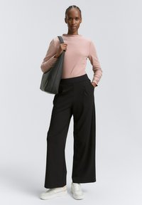 Kobieta stoi z warkoczem, ubrana w jasnoróżową bluzkę z długim rękawem, czarne szerokie spodnie, białe sneakersy i trzyma dużą czarną torbę na ramię.