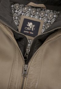Braune Lederjacke mit schwarzem Futter, ausgestattet mit zwei Reißverschlüssen, einem kompliziert gemusterten Innenfutter und einem Etikett mit einem Löwen-Logo.