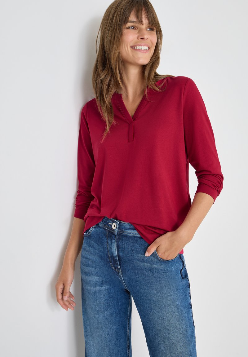 Rode longsleeve shirt met een kraag en een lichte plooidetail, gecombineerd met blauwe denim jeans, die een voorzak en een relaxte fit hebben.