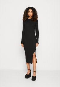 Robe midi noire à côtes avec des manches longues, fente latérale et silhouette ajustée, assortie à des talons épais noirs.
