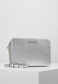 Bolso cruzado de Michael Kors en color plata, con superficie texturizada, correa de cadena y cuero, y cierre de cremallera sobre un pedestal blanco.