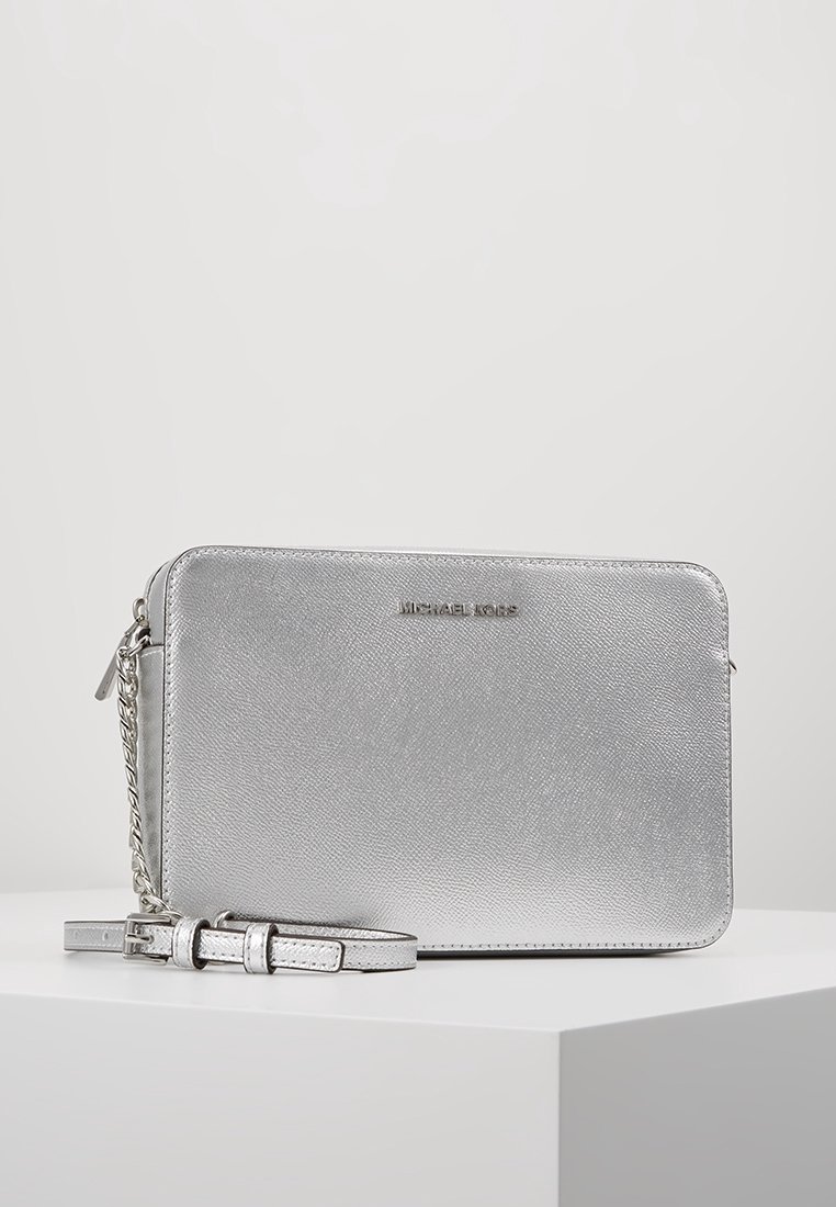 Bolso cruzado de Michael Kors en color plata, con superficie texturizada, correa de cadena y cuero, y cierre de cremallera sobre un pedestal blanco.