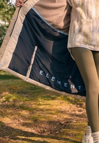 Beige ydertøj med en sort foring, der har ordet "ELEMENTS" i hvid. Indersiden har en glat tekstur og er velstruktureret.