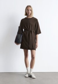 Braunes Strickkleid mit kurzen Ärmeln und einem Taillengürtel. Kombiniert mit weißen Socken und cremefarbenen Sneakers mit blauen Streifen. Schlichte, lockere Passform.