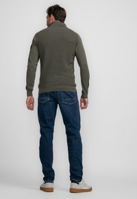 Dunkelgrüner, gerippter Pullover mit Stehkragen, kombiniert mit blauen Jeans und hellen Sneakers. Merkmale sind lange Ärmel und ein taillierter Schnitt.