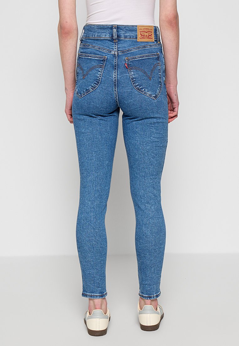 Jeans de mezclilla azul de cintura alta con una silueta ajustada, bolsillos traseros distintivos con un diseño bordado y una etiqueta de cuero con la marca.