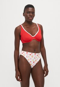 Rød ribbet bikini topp med hvit kant og v-hals design, parret med høytaljede hvite truser med et fargerikt fruktmønster.