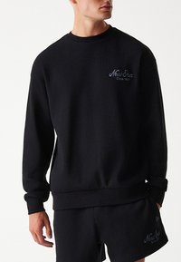 Uomo che indossa una felpa a manica lunga nera abbinata a pantaloncini con il logo "New Era Since 1920" sul petto e sulla gamba.