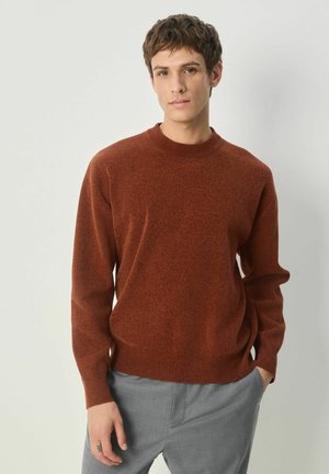 Junger Mann in einem rostfarbenen Pullover und grauen Hosen, der mit Händen in den Taschen vor einem hellen neutralen Hintergrund steht.