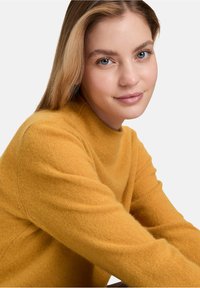 Pull en maille jaune avec un col montant, texture douce et manches longues. Le tissu semble chaud et confortable. Design simple sans motifs.