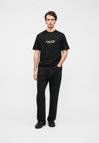 Μαύρο κοντομάνικο μπλουζάκι με χρυσό κείμενο και γραφικό, συνοδευόμενο από άνετο μαύρο τζιν παντελόνι και μαύρα παπούτσια. Απλό λευκό φόντο.