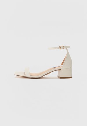 Beige block-heeled sandal with a square toe. Features a slim ankle strap with a buckle, smooth leather-like texture, and a moderate heel height.