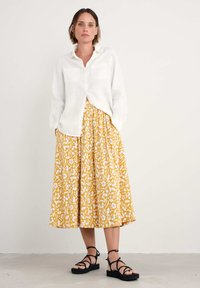 Vit linneskjorta med knappar och två fickor, kombinerad med en gul blomstermönstrad midikjol och svarta remsandaler, på en neutral bakgrund.