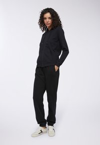 Felpa nera con zip, cappuccio e tasche frontali, abbinata a pantaloni jogger neri. Sneakers bianche con un accento nero. Texture liscia.