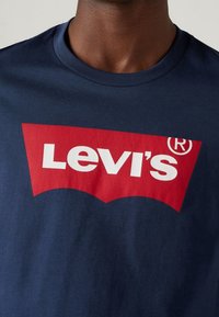 Námořnická modrá bavlněná tričko s velkým červeným logem Levi's s bílým písmem, umístěné na texturovaném pozadí látky.