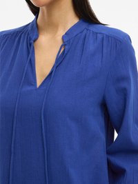 Plava bluza dugih rukava s mandarin ovratnikom, mekane teksture tkanine i nabora blizu izreza. Sadrži suptilne detalje s vezanjem na prednjem dijelu.