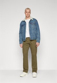 Jeansjacke mit Lammfellkragen, hellblaue Farbe, mit Knopfverschluss. Weißes T-Shirt darunter. Olivgrüne Cargo-Hosen, weiße Sneakers.