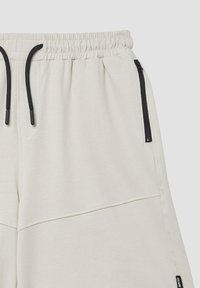 Helle Creme Sweatpants mit elastischem Bund, ausgestattet mit schwarzen Kordeln und Reißverschlusstaschen an den Seiten. Glatte Textur mit Nahtdetails.